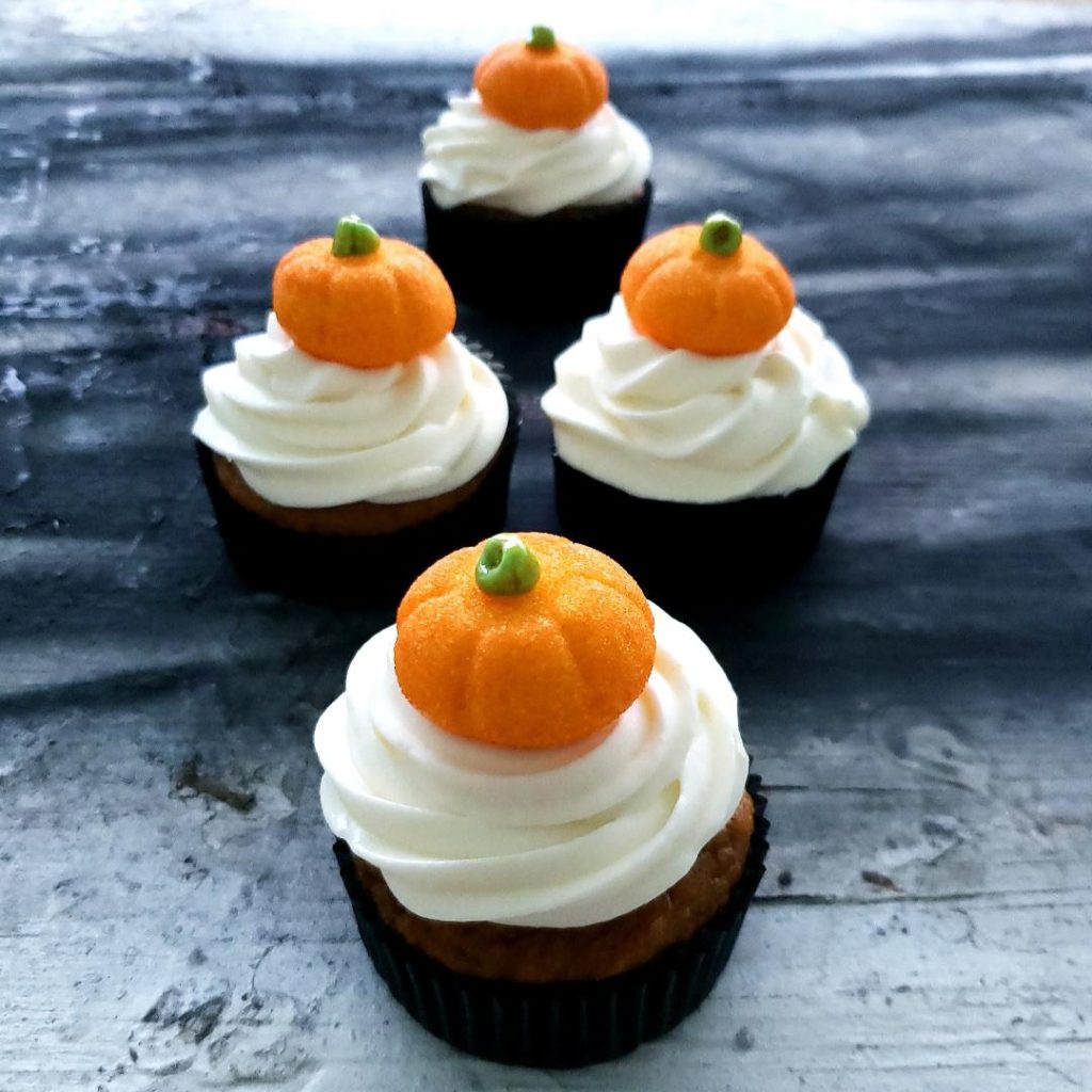 functional image pumpkin patch cupcakes side view on distressed gray and black background  4 cupcakes in a formation one in back to side by side in the middle and one in front all have black cupcake liners white cream cheese icing and a glittery orange pumpkin shaped marshmallow on top