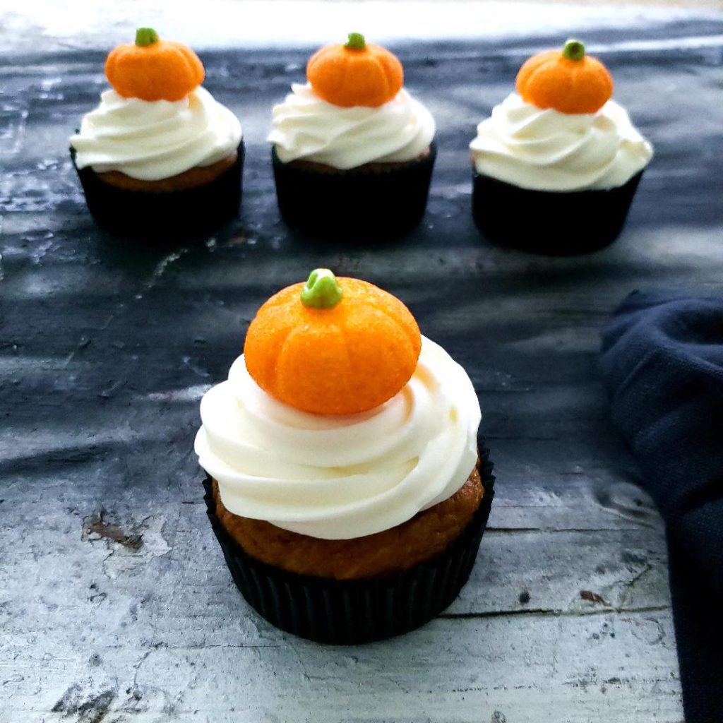 functional image pumpkin patch cupcakes side view on distressed gray and black background three cupcakes lined up in the back and one in the front all have black cupcake liners white cream cheese icing and a glittery orange pumpkin shaped marshmallow on top