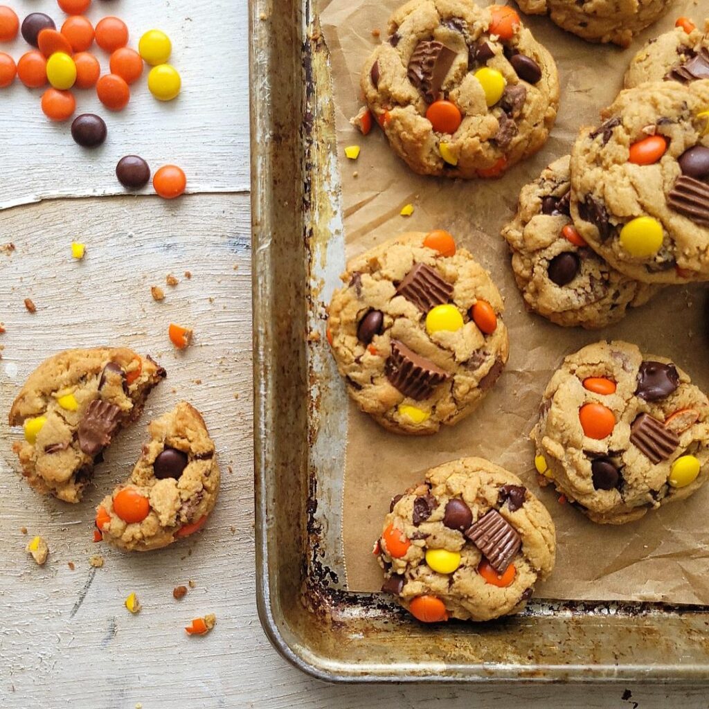 reese's pieces cookies. top down view of baked cookies in a random pattern on a cookie sheet. to the left there are reese's pieces candies and a broken cookie. 
