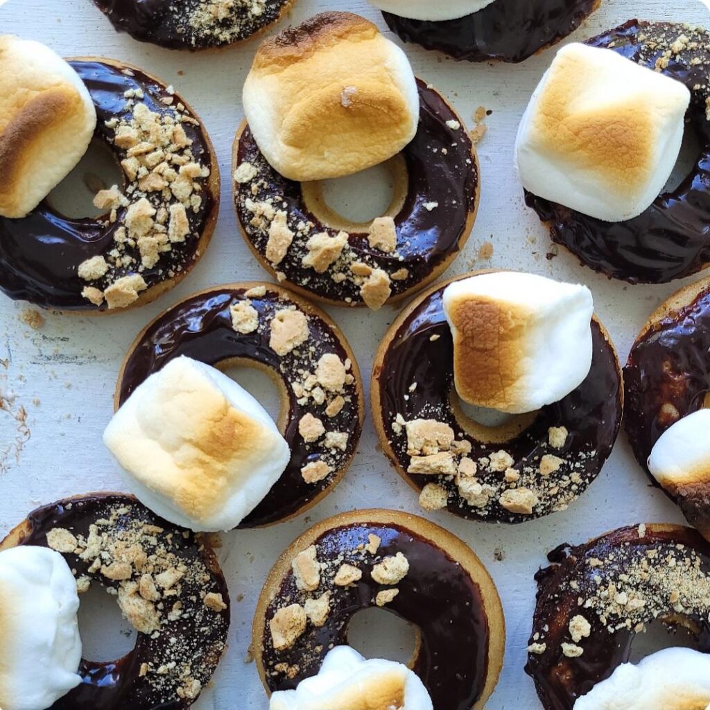 s'mores donuts. top down view of baked donuts topped with chocolate ganache, toasted marshmallows and graham cracker crumbs.