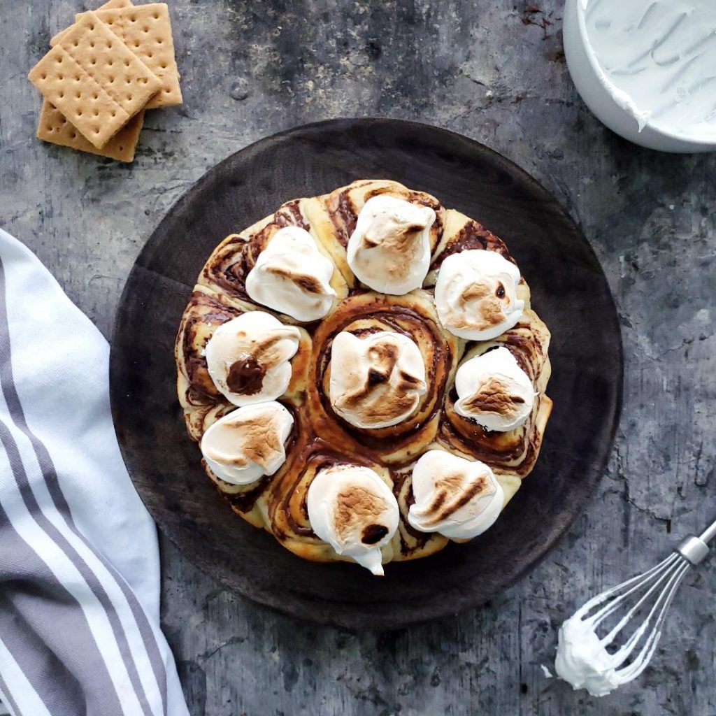 s'mores cinnamon rolls with marshmallow meringue top down photo distressed gray table   in the center of the photo is a round black charger plate with the full batch of smores rolls with toaster marshmallow meringue on top.  top left is a white bowl of marshmallow meringue bottom left is a gray and white striped linen and bottom right a metal whisk with marshmallow meringue top left is three graham crackers