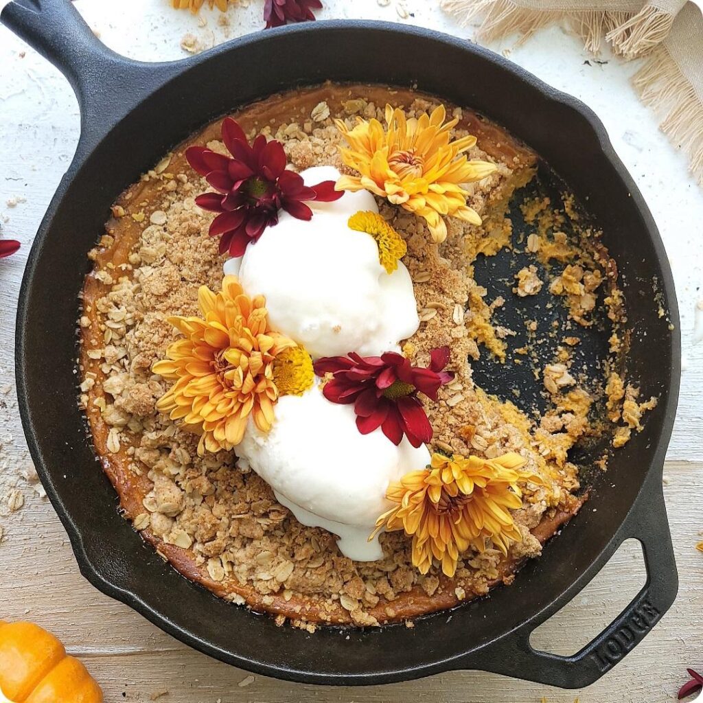 pumpkin crisp baked in a cast iron skillet. top down view of crisp garnished with scoops of vanilla ice cream and fall flowers. a section of the crisp has been scooped away and a few crumbs remain.