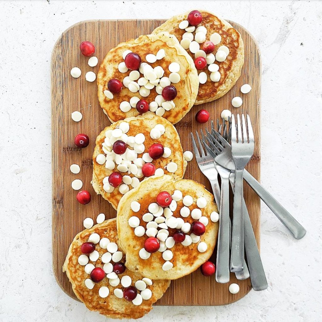 functional image white chocolate chip pancakes 4 laid out on a wooden cutting board with four forks garnished with white chocolate chips and fresh cranberries