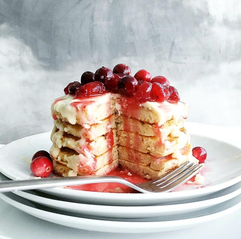 functional image white chocolate chip pancakes stacked on a plate with melted white chocolate drizzling down the sides and fresh cranberries piled on top pancakes have a wedge cut out like a cake so you can see the layers