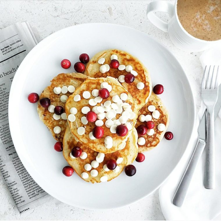 functional image white chocolate chip pancakes top down image on a plate with white chocolate chips and fresh cranberries a coffee mug and a newspaper