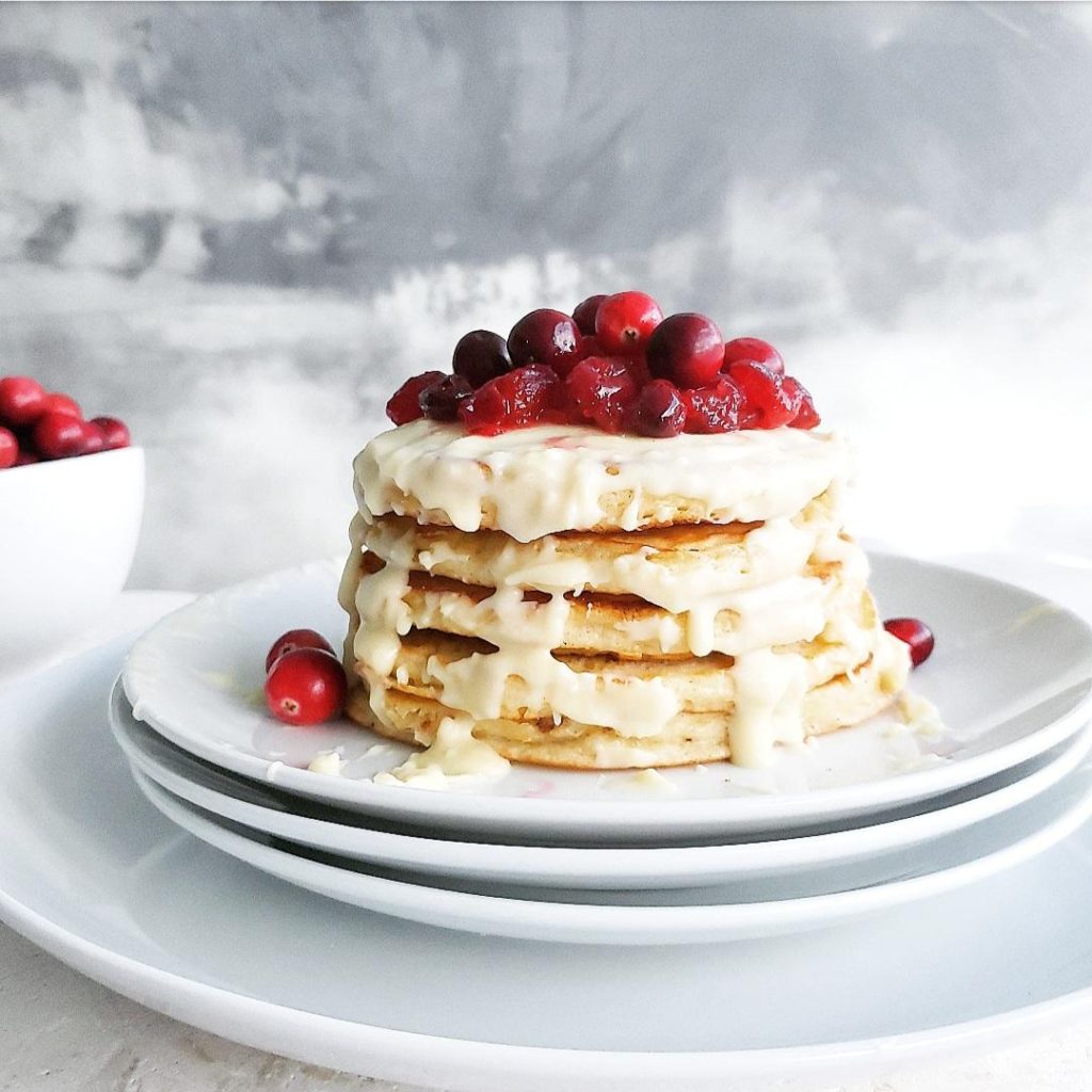 functional image white chocolate chip pancakes stacked on a plate with melted white chocolate drizzling down the sides and fresh cranberries piled on top
