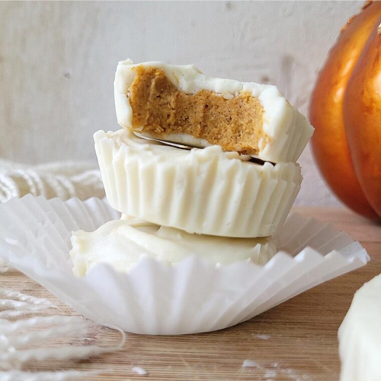 white chocolate pumpkin cups. three stack in a white cupcake liner. with an orange pumpkin in the background. top pumpkin cup has been bitten.
