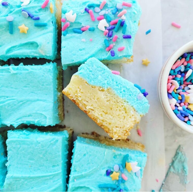frosted sugar cookie bars. top down view of bars cut into squares and frosted with buttercream frosted tinted blue. one cookie is on its side so you can see the inner crumb.