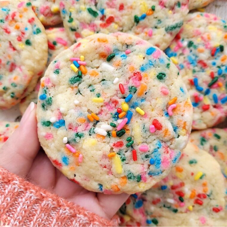 funfetti cookies. close up view of a hand holding a large sugar cookie with rainbow sprinkles. the background is a pile of more sprinkle cookies.