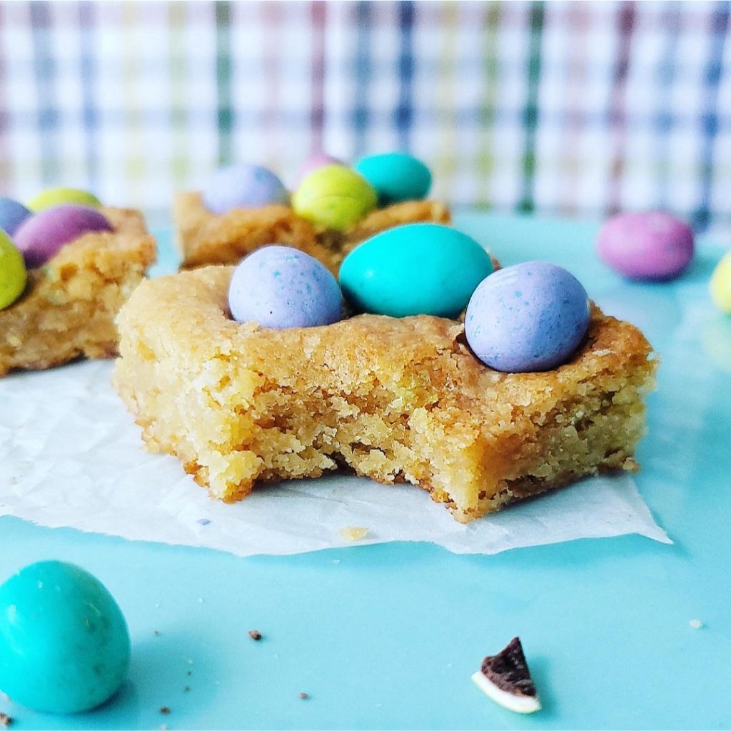  m&m easter blondies. close of view of an easter blondie topped with pastel colored m&m eggs. one bar has a bite missing