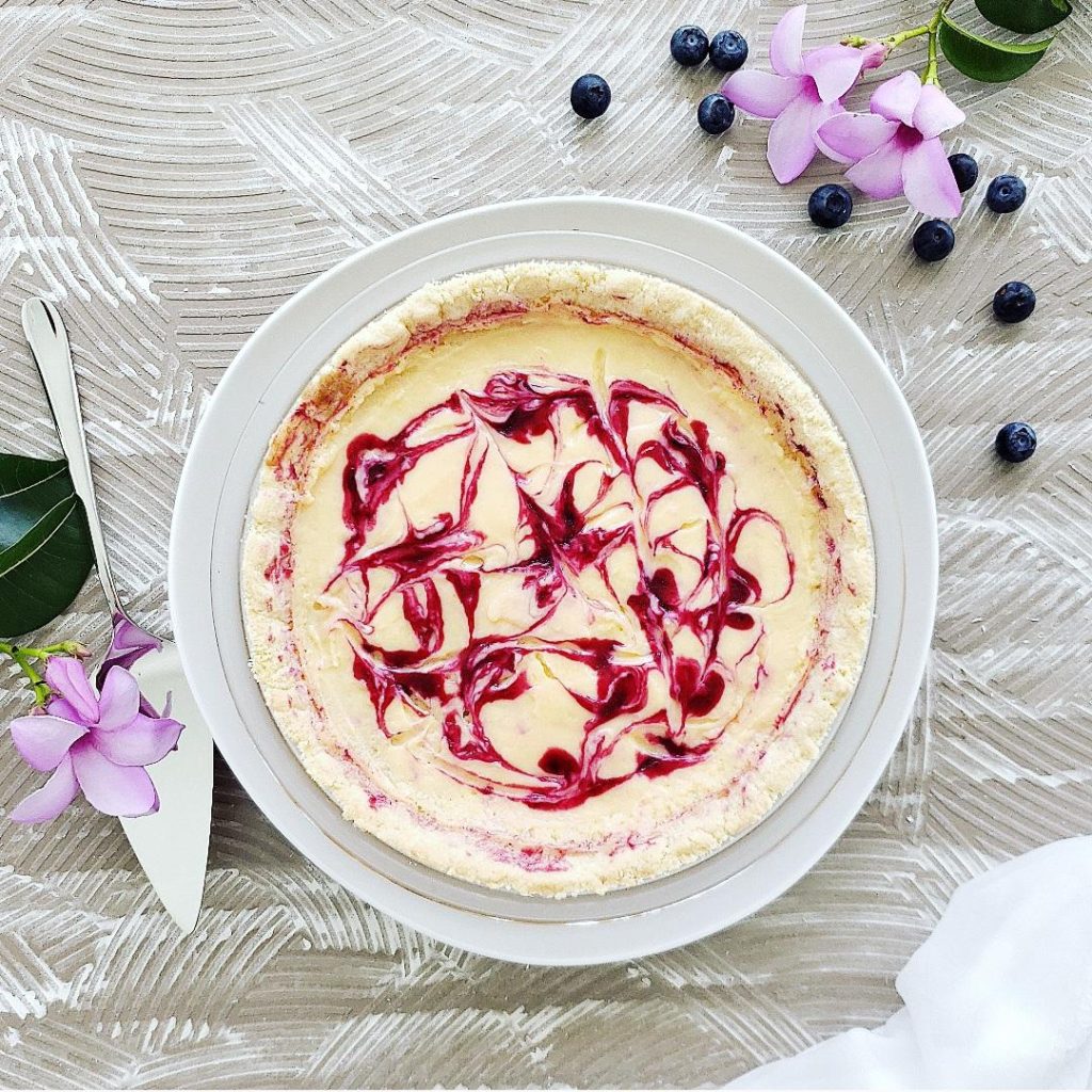 top down view of lemon pie swirled with blueberry sauce. background is styled with fresh blueberries purple flowers and a sterling silver pie server lemon blueberry pie recipe