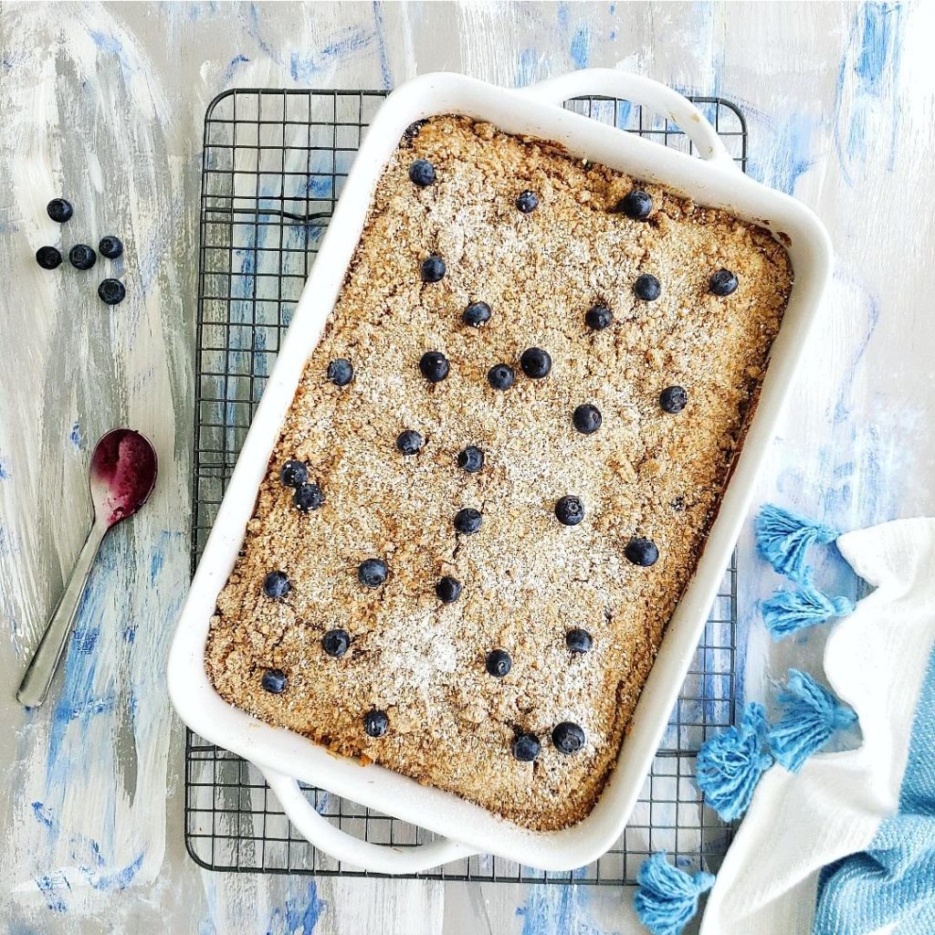 sour cream blueberry coffee cake. top down view of baked  cake with a crumble topping in a 9 x 13 pan garnished with fresh blueberries