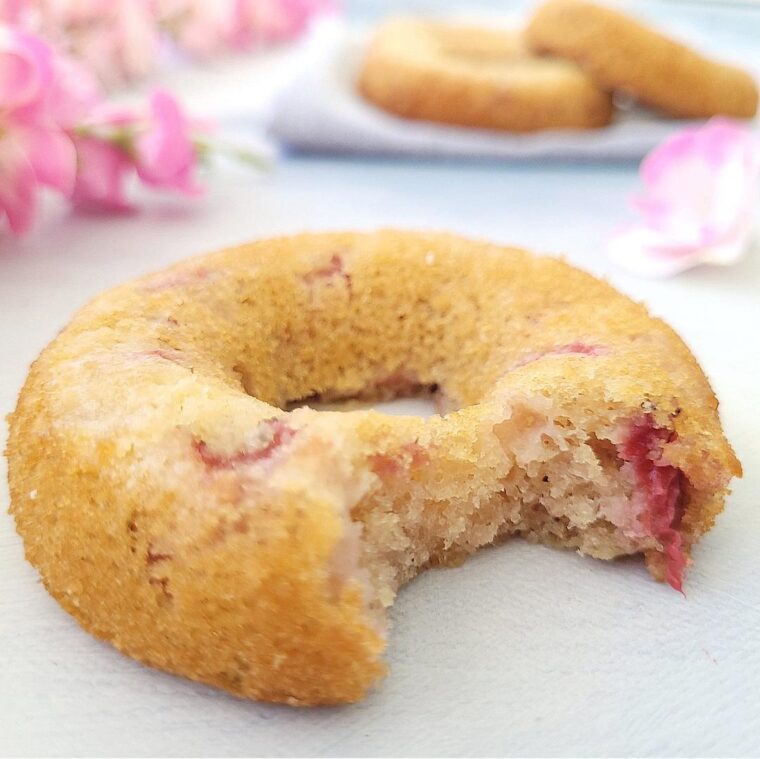 close up view of a baked strawberry donut with a bite taken out of it so you can see the fluffy crumb and bits of strawberries inside. background has pink flowers and two more donuts.