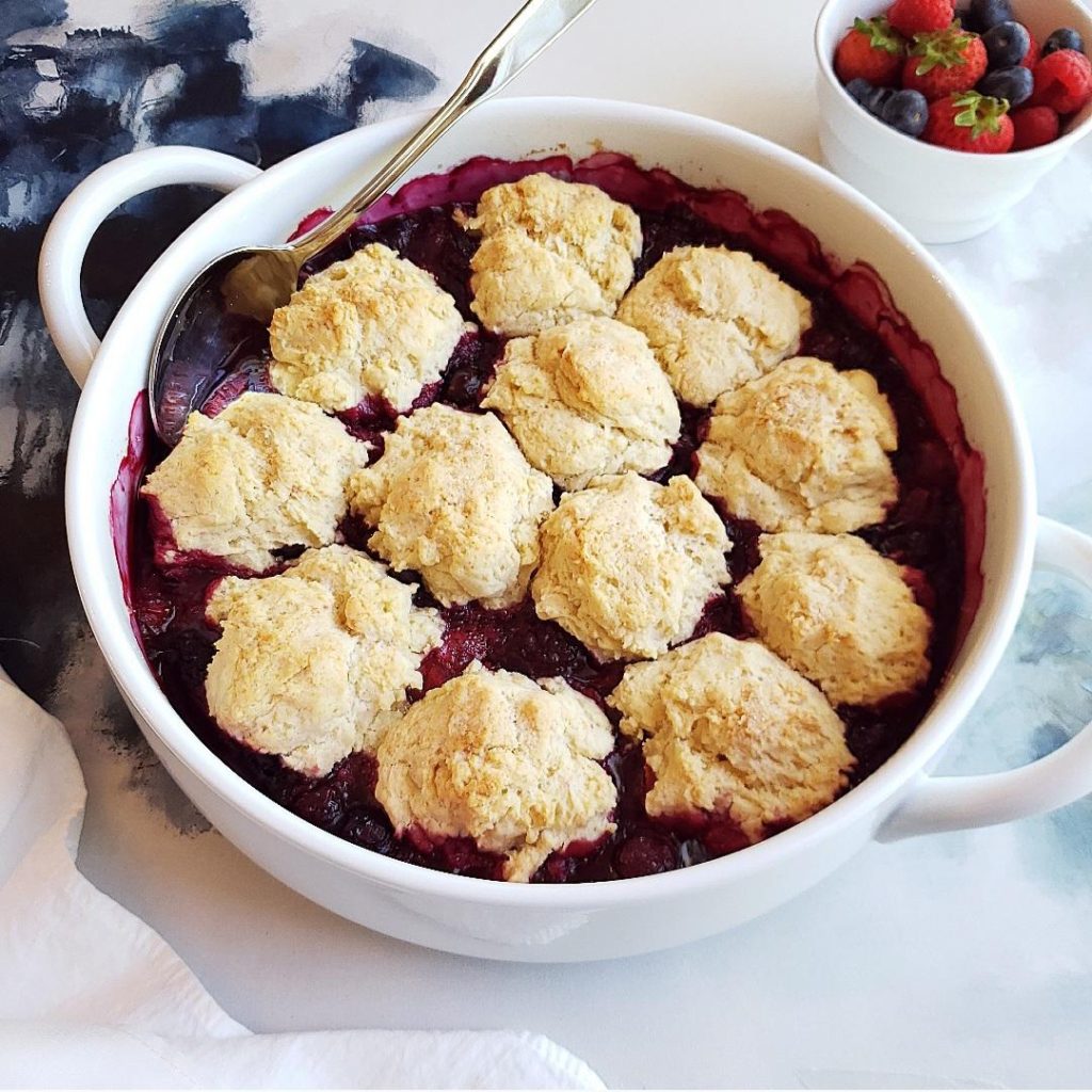 functional image mixed berry cobbler buttermilk biscuits in a white baking dish with a bowl of fresh fruit