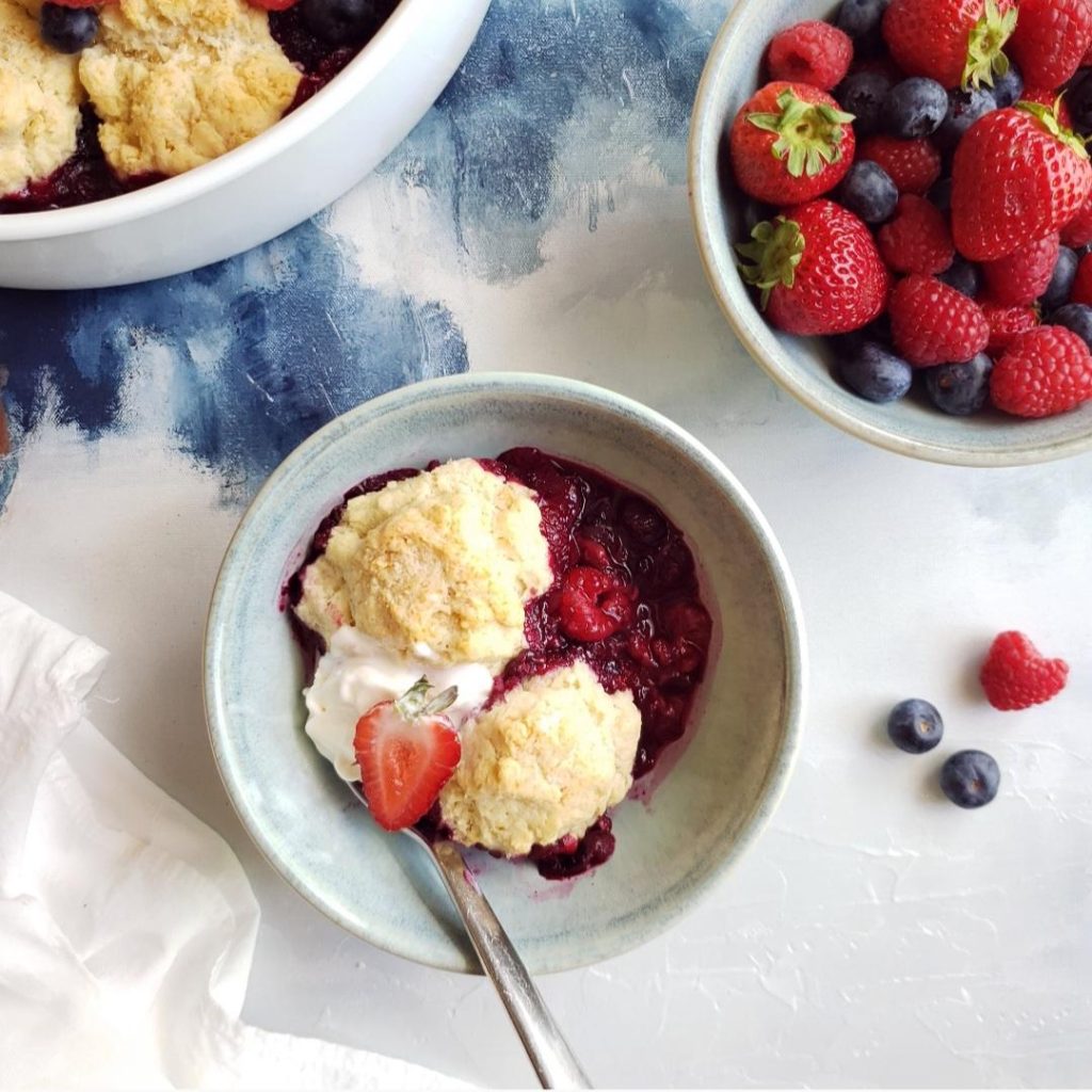 functional image mixed berry cobbler in a pale blue bowl with a spoon two biscuits and melted ice cream