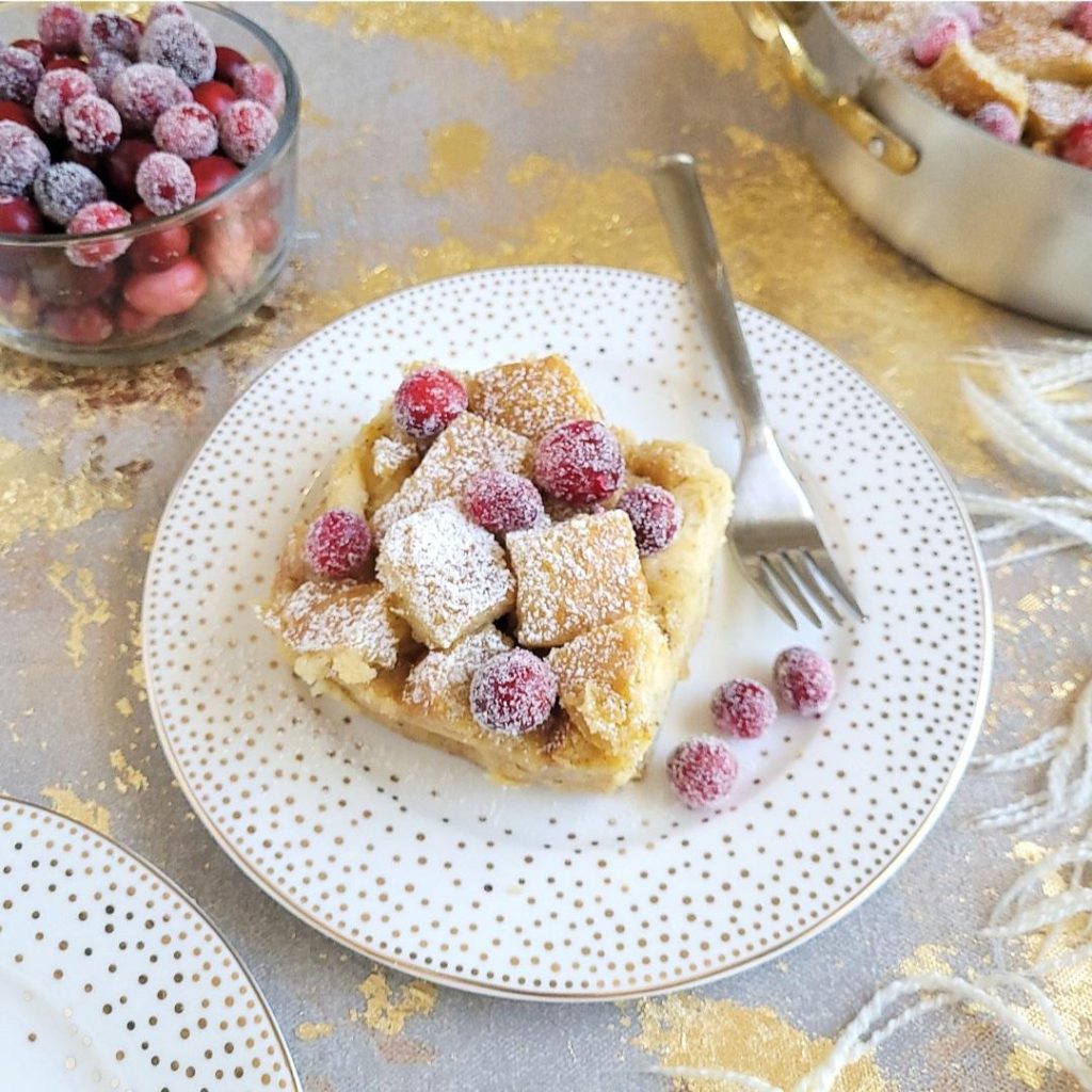 functional image eggnog bread pudding square slice on a round plate with gold polka dots. dusted with powdered sugar and garnished with sugared cranberries