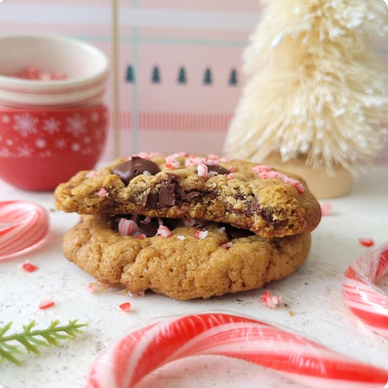 peppermint chocolate chip cookies. side view of two cookies stacked. top cookie has a bite missing and is topped with chocolate chips and broken bits of candy cane. background is a white bottlebrush tree, small red bowls with snowflakes and a pink backdrop with tiny green christmas trees.