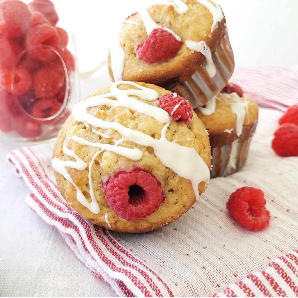 white chocolate raspberry muffins drizzled with melted white chocolate three muffins two stacked in back one tilted forward in front with fresh raspberries