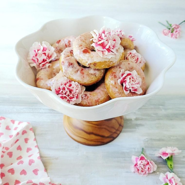functional image cherry donuts with cherry vanilla glaze piled on a white cake stand with a wooden base and garnished with pink carnations