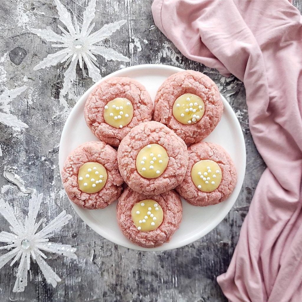 functional image pink velvet thumbprint cookies with cream cheese filling and white sprinkles top down on a white plate with crystal snowflakes and a pink linen