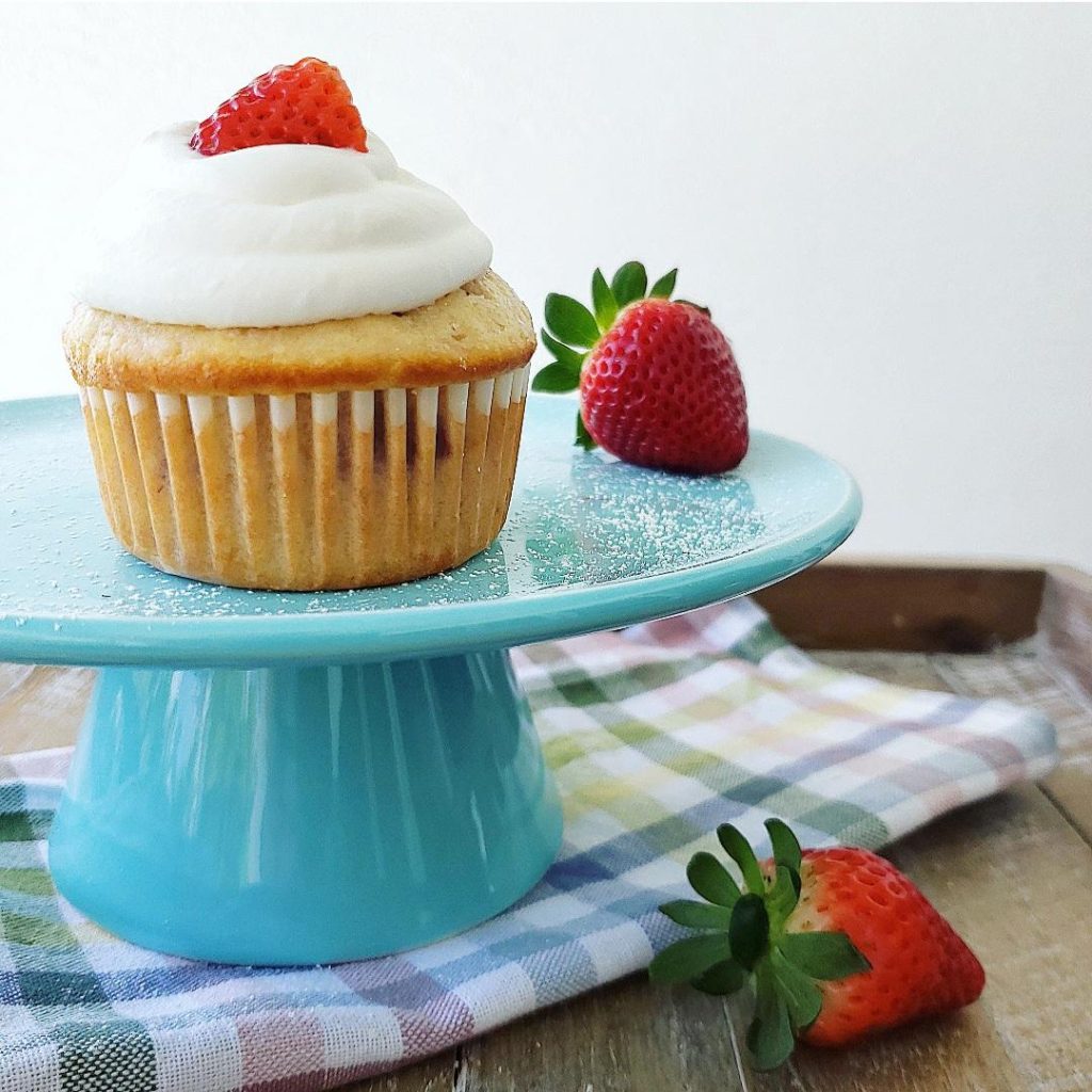 functional image strawberry shortcake muffins on a blue cake stand with whipped cream and a fresh strawberry on top of a muffin