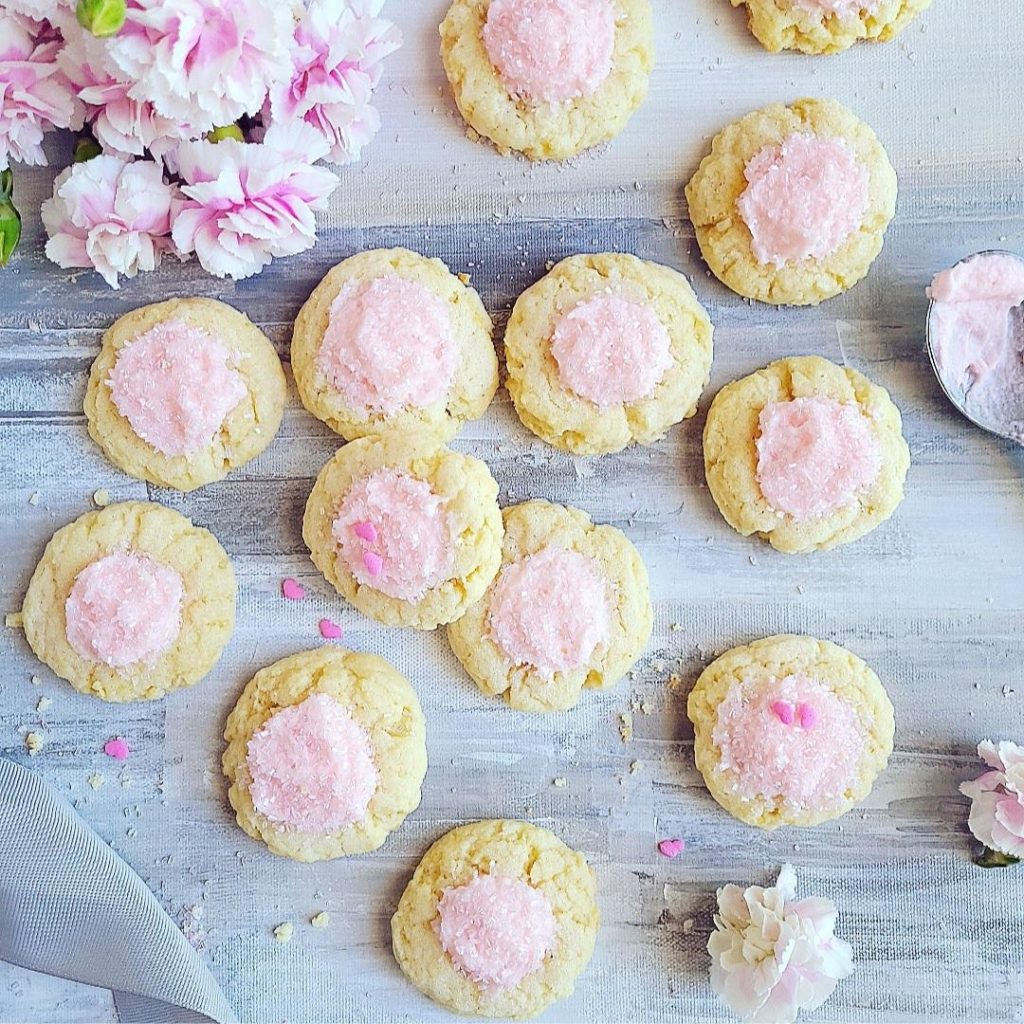 thumbprint cookies with icing pink icing with pink sprinkles top down 1 dozen cookies scattered with crumbs, a spoon with pink icing and pink carnations