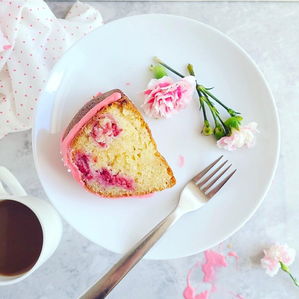 functional image white chocolate raspberry bundt cake one slice on its side on a white plate with pink carnations and a fork you can see the moist vanilla crumbs and juicy raspberries in the cake