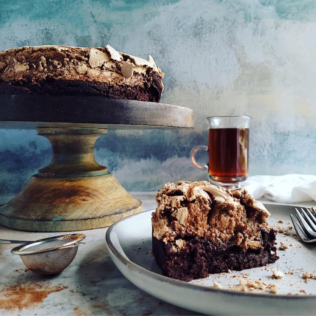 functional image brownie cake with chocolate meringue on a wooden cake stand blue background cut slice on a plate in foreground. coffee in glass cup in background