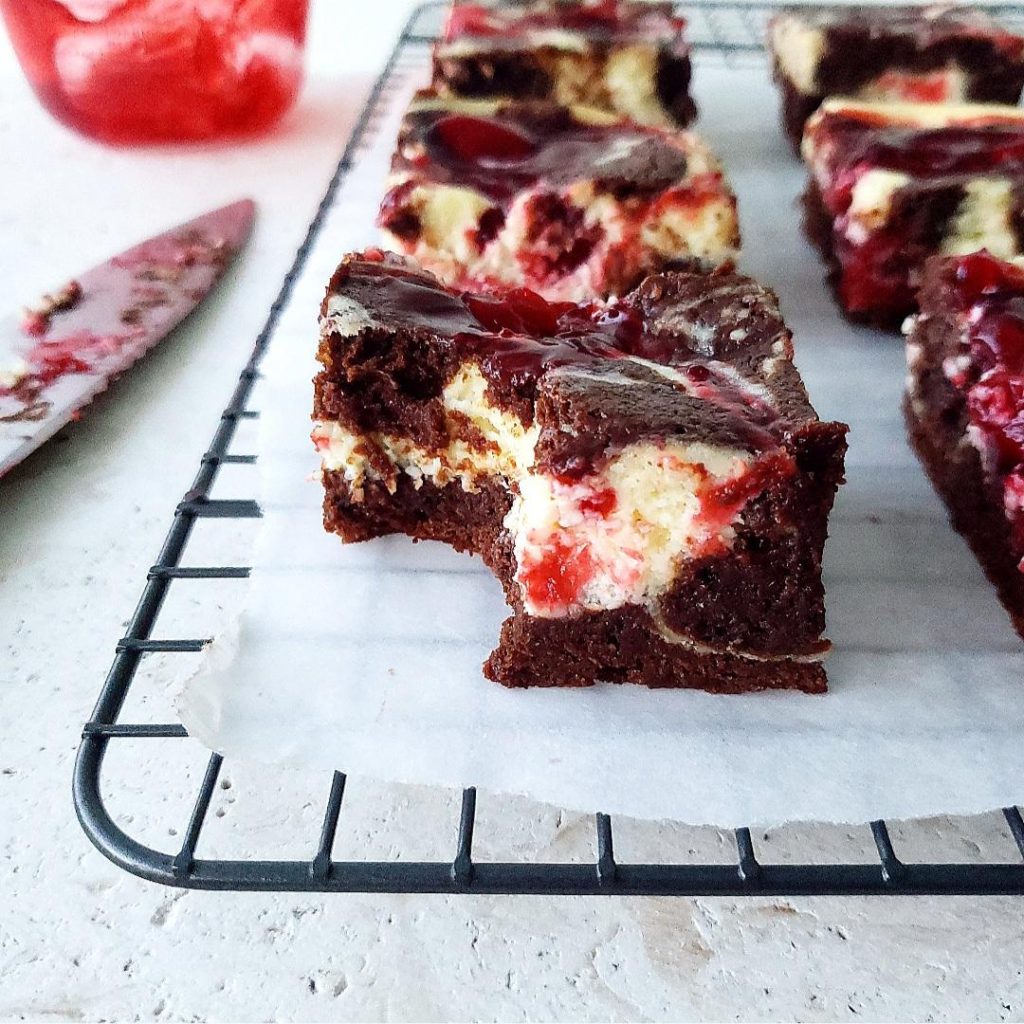 functional image cherry cheesecake brownies cut into squares on a wire baking rack with a bite missing from one piece. styled with a messy knife and a jar of cherries in the background