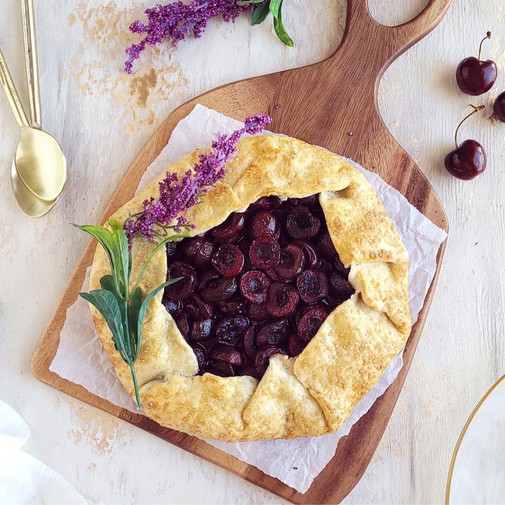 functional image cherry galette top down uncut on a wooden cutting board styled with lavender two gold spoons and 3 fresh cherries with stems