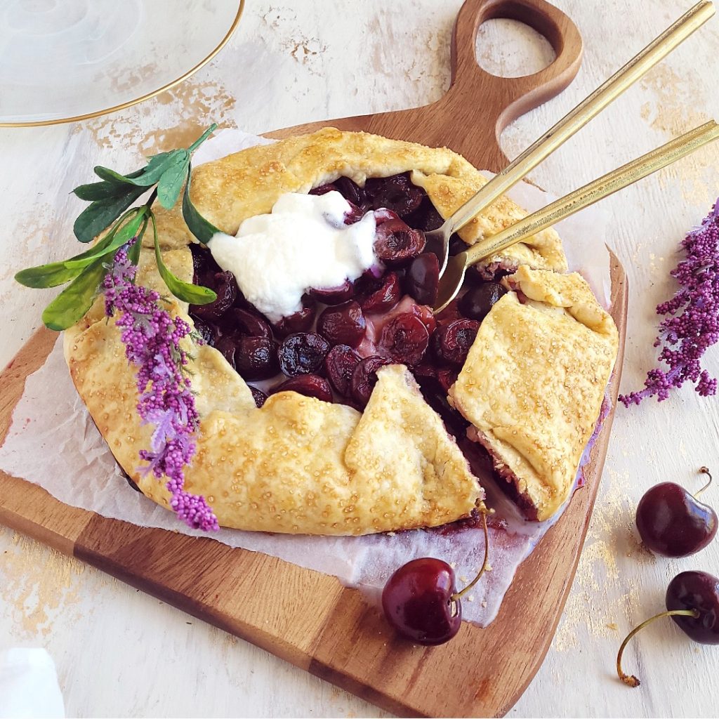 functional image cherry galette on a wooden cutting board with a melty scoop of vanilla ice cream and two gold spoons in the center of the pie