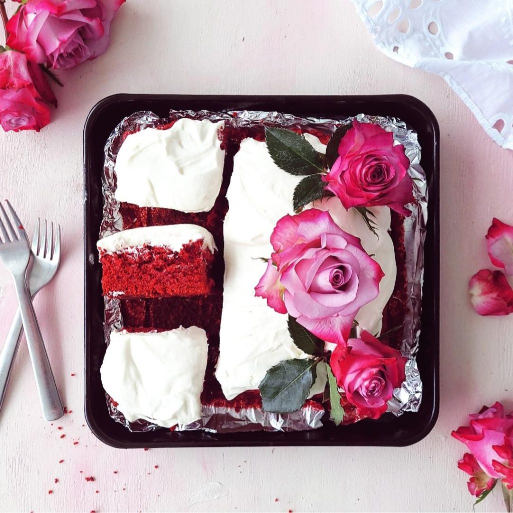 red velvet brownies with cream cheese frosting and purple roses three slices cut one on its side so you can see the red crumb styled with forks and fresh flowers on a pick background