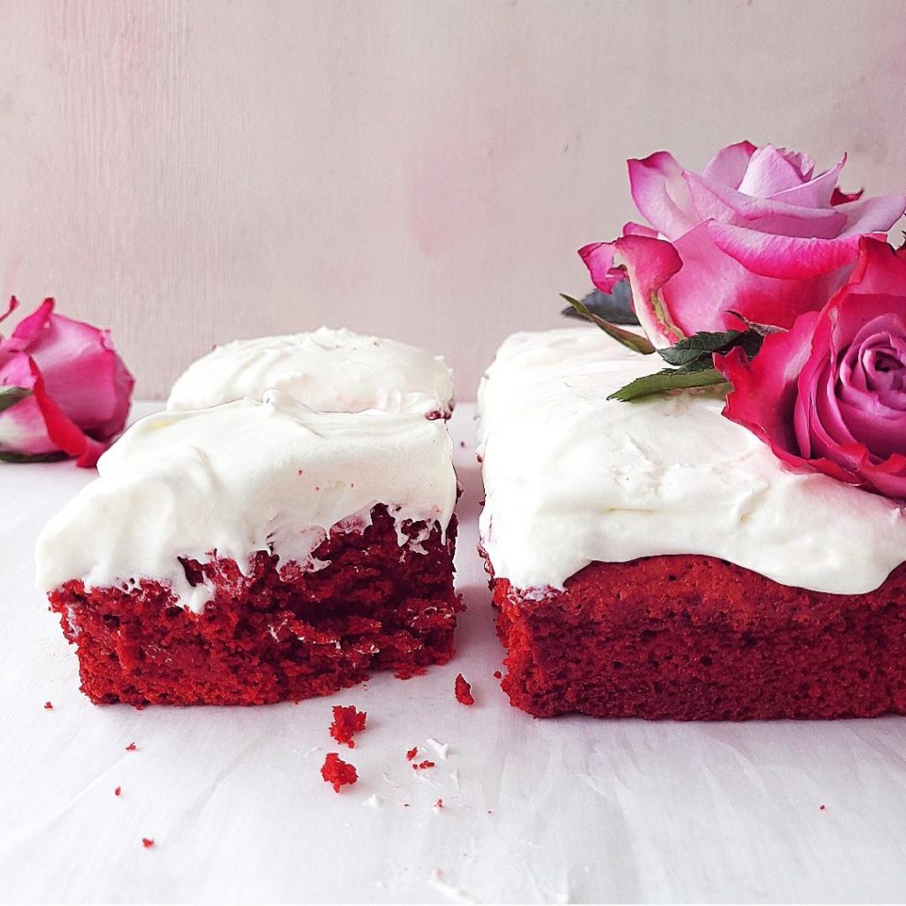 functional image red velvet brownies with cream cheese frosting side view with a pink background styled with pink and purple fresh roses one slice has a bite missing and red crumbs