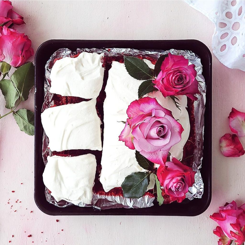 functional image red velvet brownies top down on a pink background. brownies are in a square black tray with three slices cut and the uncut portion is styled with pink and purple fresh roses and greenery. frosted with cream cheese icing. 