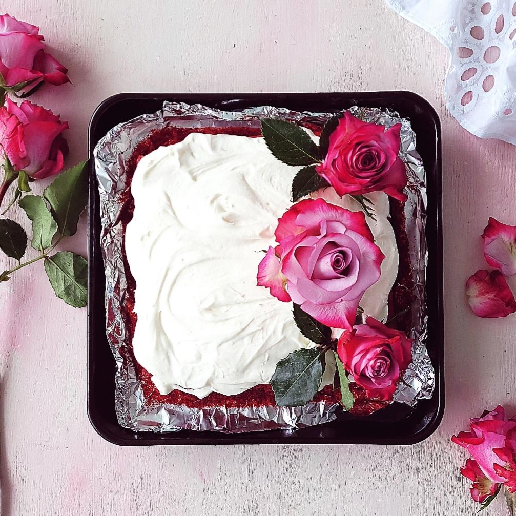 functional image red velvet brownies frosted with cream cheese icing top down uncut in a black tray lined with aluminum foil the right side of the brownies is styled with fresh roses