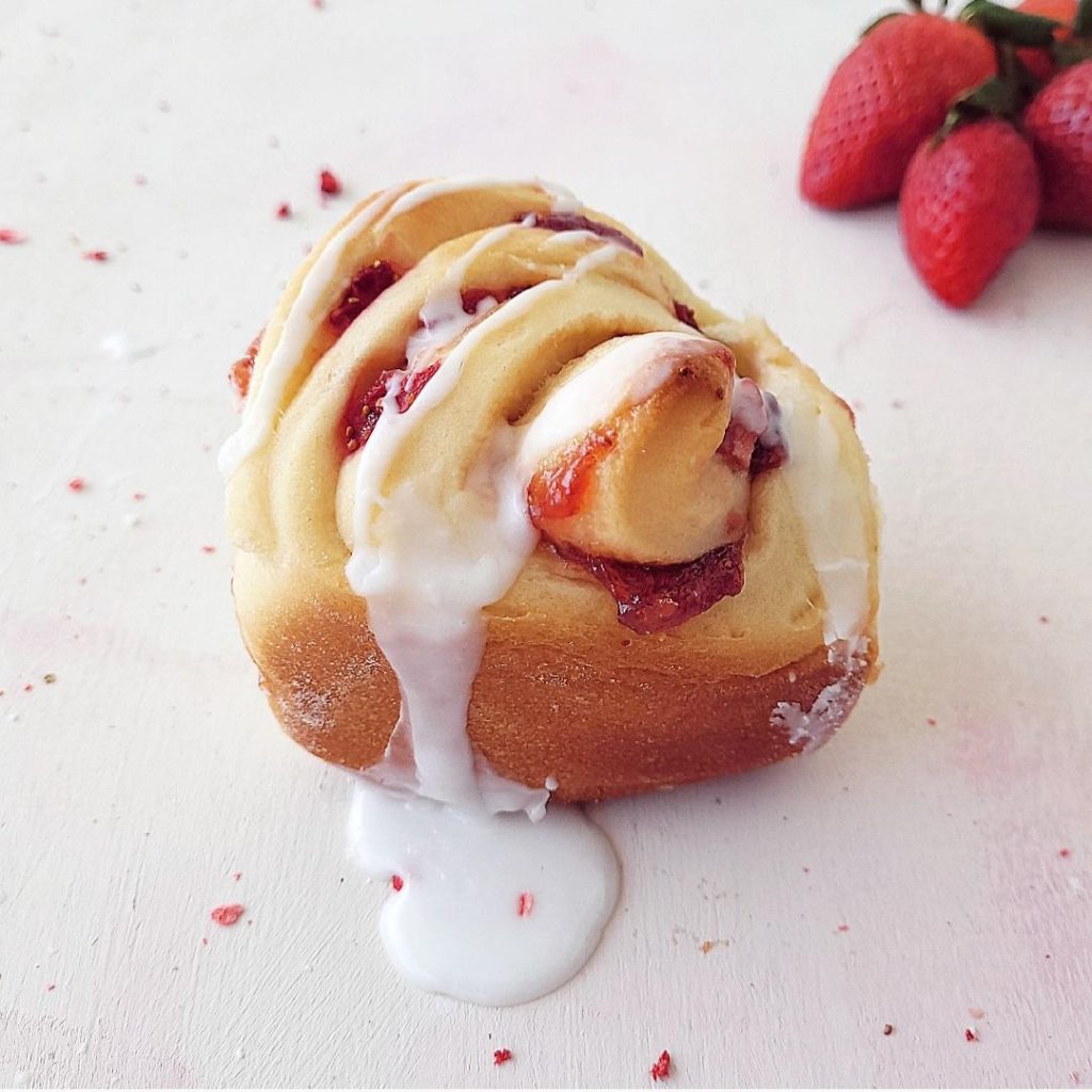 functional image close up of strawberry cinnamon rolls single roll with white icing dripping down the sides and strawberries in the background