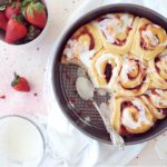 functional image strawberry cinnamon rolls top down photo with icing in a metal baking pan two rollls have been removed styled with fresh strawberries and a bowl of icing and a spoon covered in icing