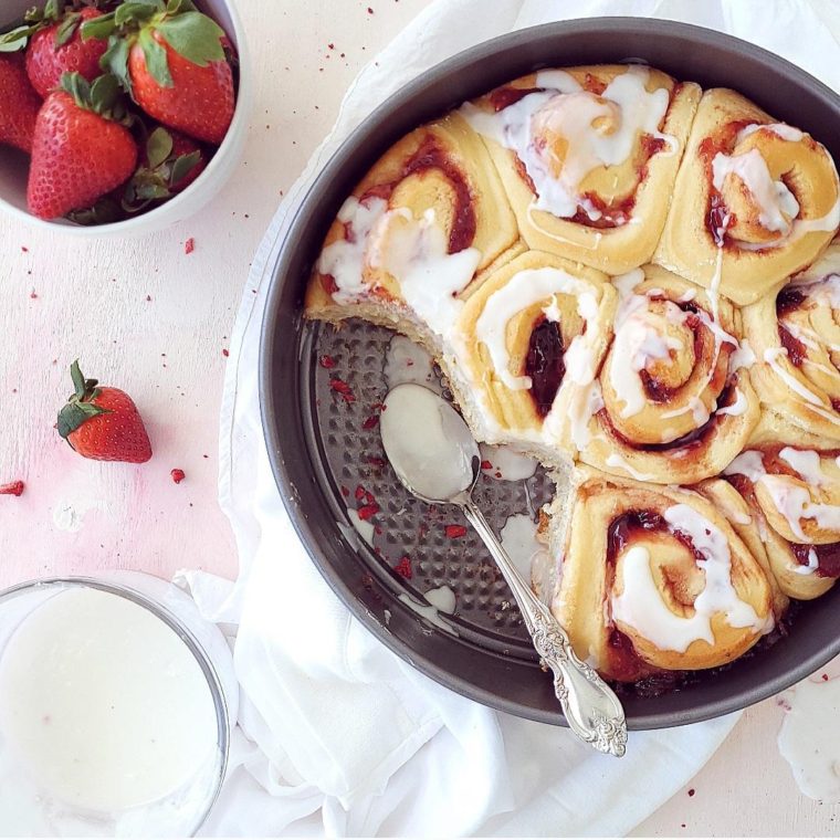 functional image strawberry cinnamon rolls top down photo with icing in a metal baking pan two rollls have been removed styled with fresh strawberries and a bowl of icing and a spoon covered in icing