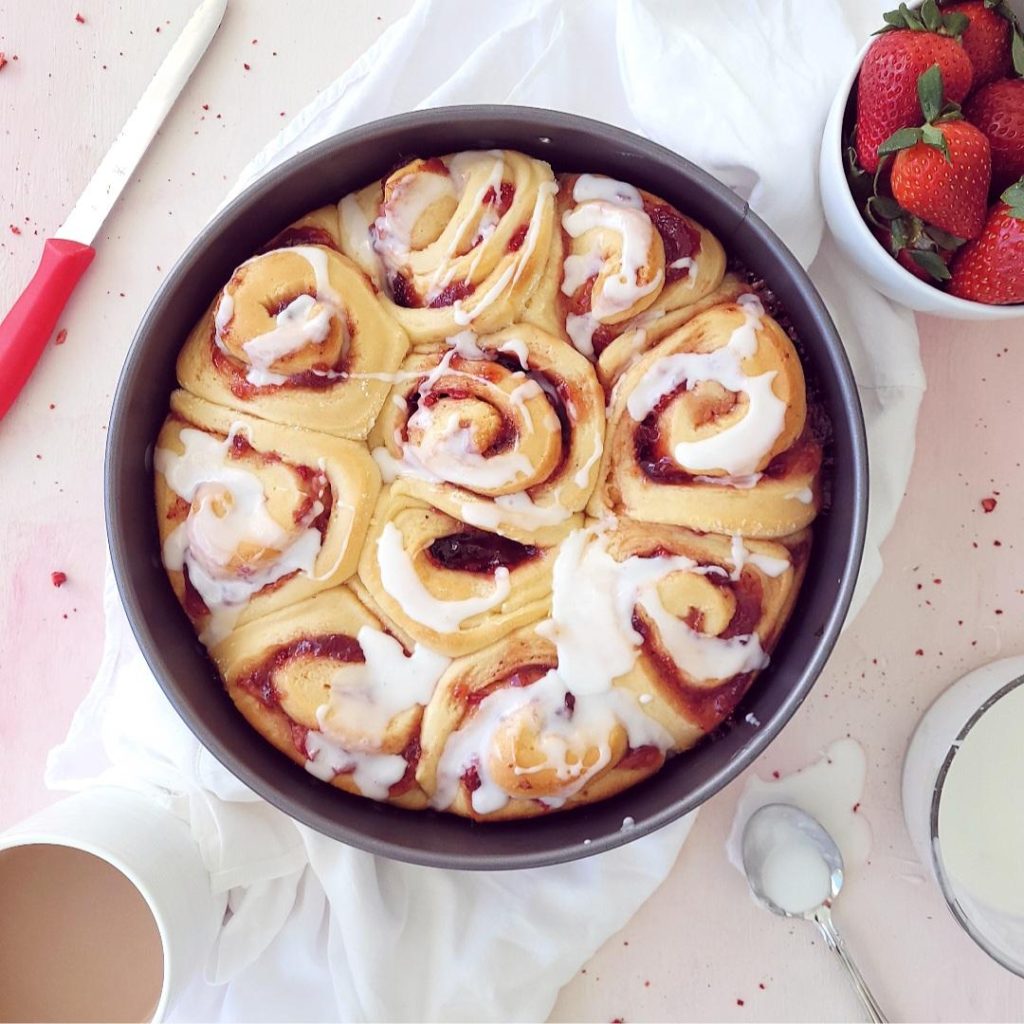 functional image strawberry cinnamon rolls top down with icing uncut in a metal baking pan with a bowl of strawberries and a red handled knife