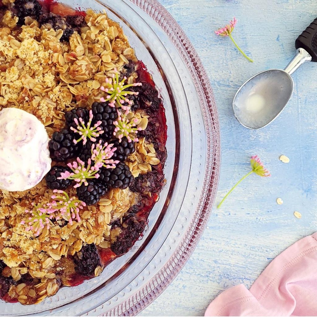functional image blackberry crisp rule of thirds cropping with fresh blackberries and a scoop of ice cream on a pink glass cake stand with a used ice cream scoop and tiny pink flowers