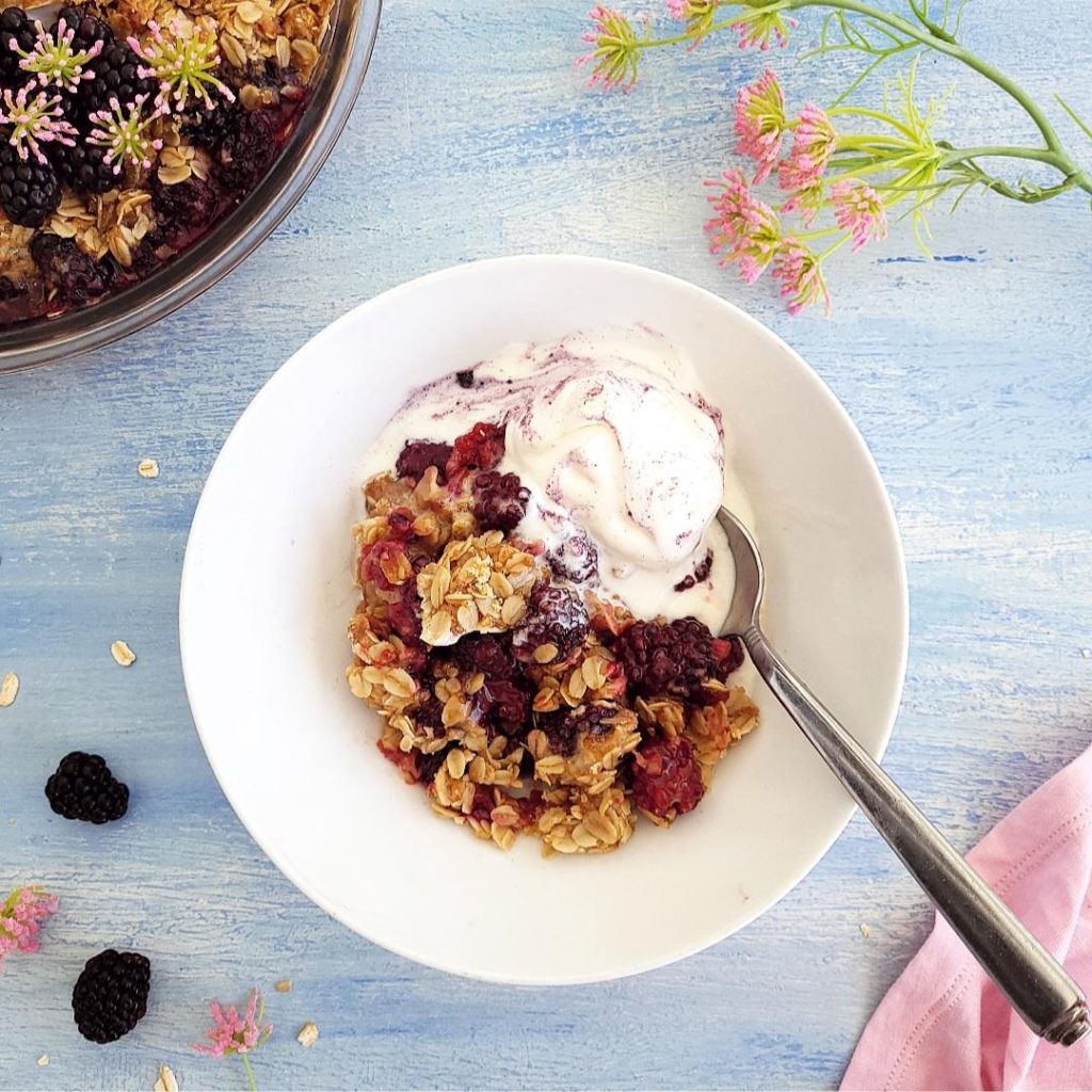 functional image blackberry crisp scooped into a white bowl with vanilla ice cream and a spoon on a blue background