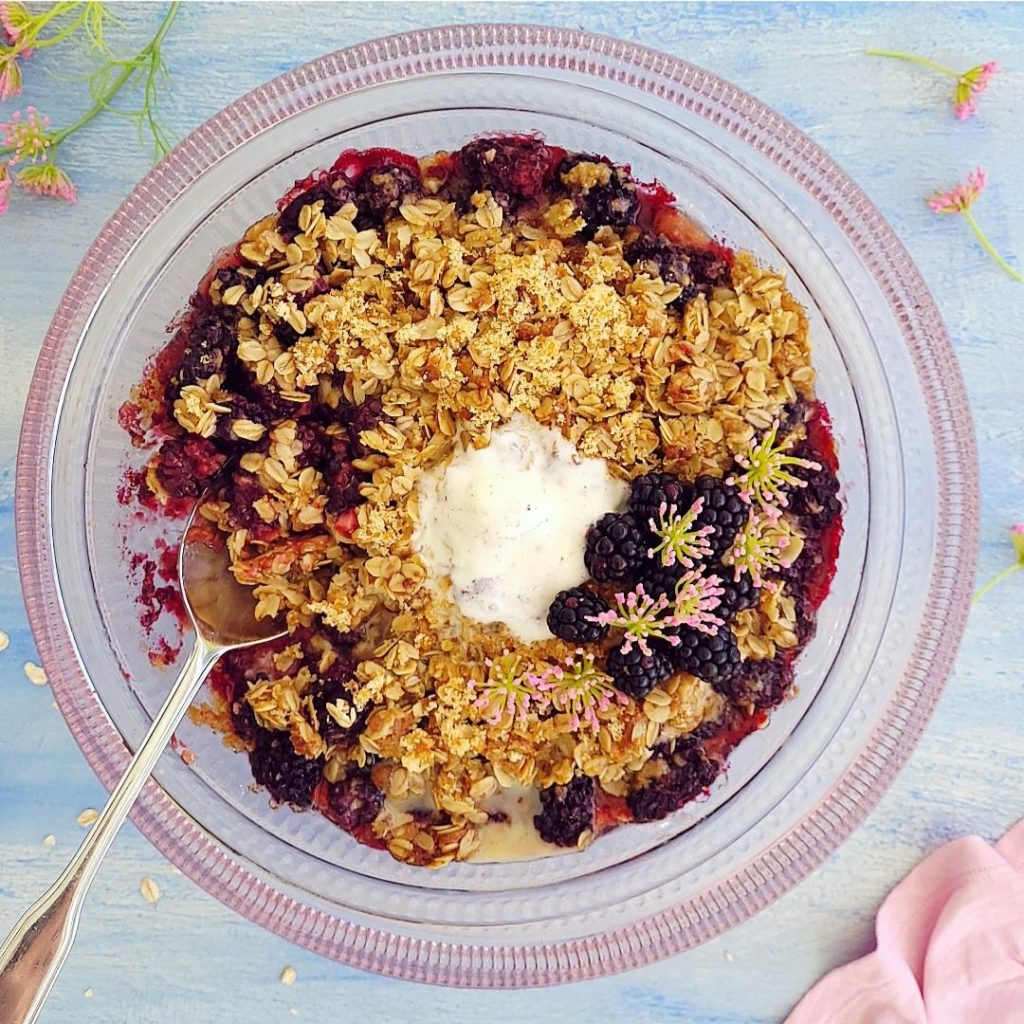 functional image blackberry crisp top down on a pink glass cake stand with fresh blackberries a melted scoop of ice cream and a spoon on a blue background