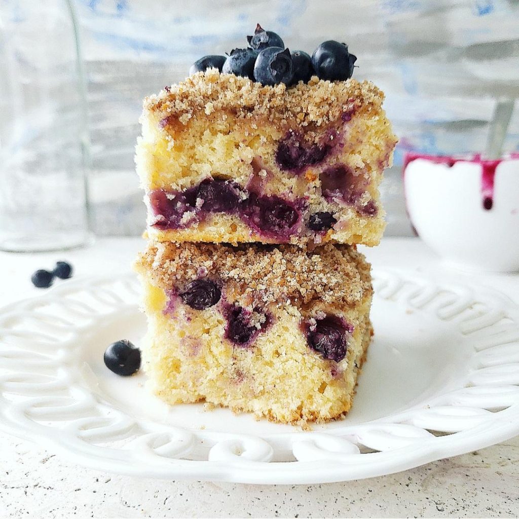 functional image blueberry coffee cake with sour cream side view of  two square slices stacked on a white scalloped plate. close up of crumbs and berries in cake blue background