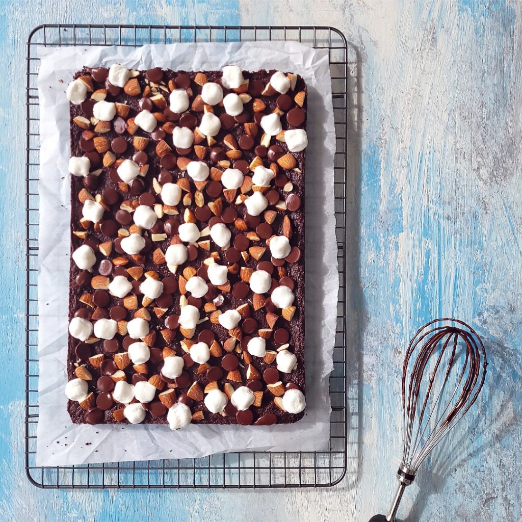 functional image rocky road brownies top down uncut on parchment on a baking rack on a distressed blue background with a chocolaty whisk to the side