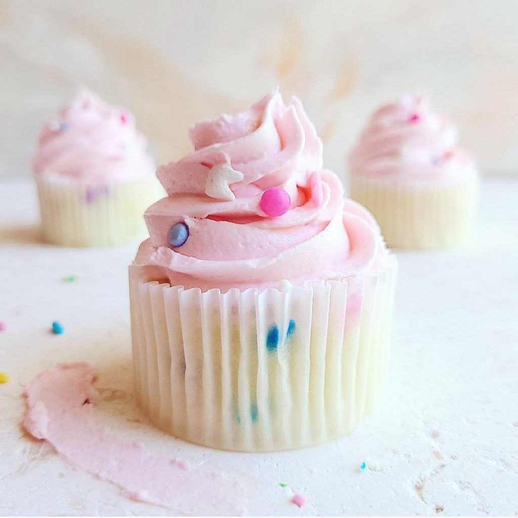 functional image sprinkle cupcakes close of of one with two in the background iced with pink frosting and garnished with pink blue and white spinkles
