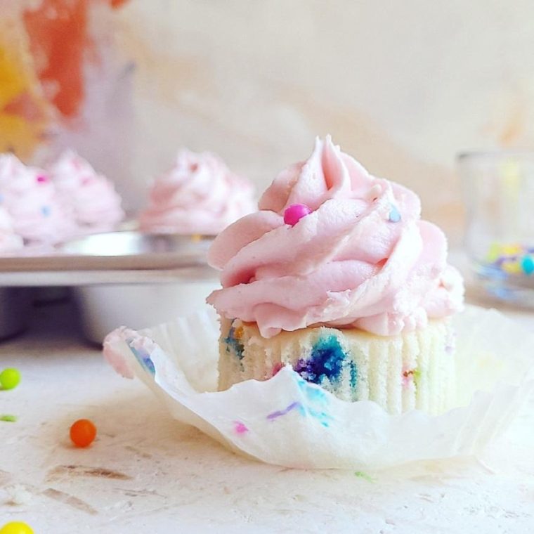 confetti sprinkle cupcakes with pink frosting wrapper removed to see white cake crumb and sprinkles baked in shiny cupcake pan in background