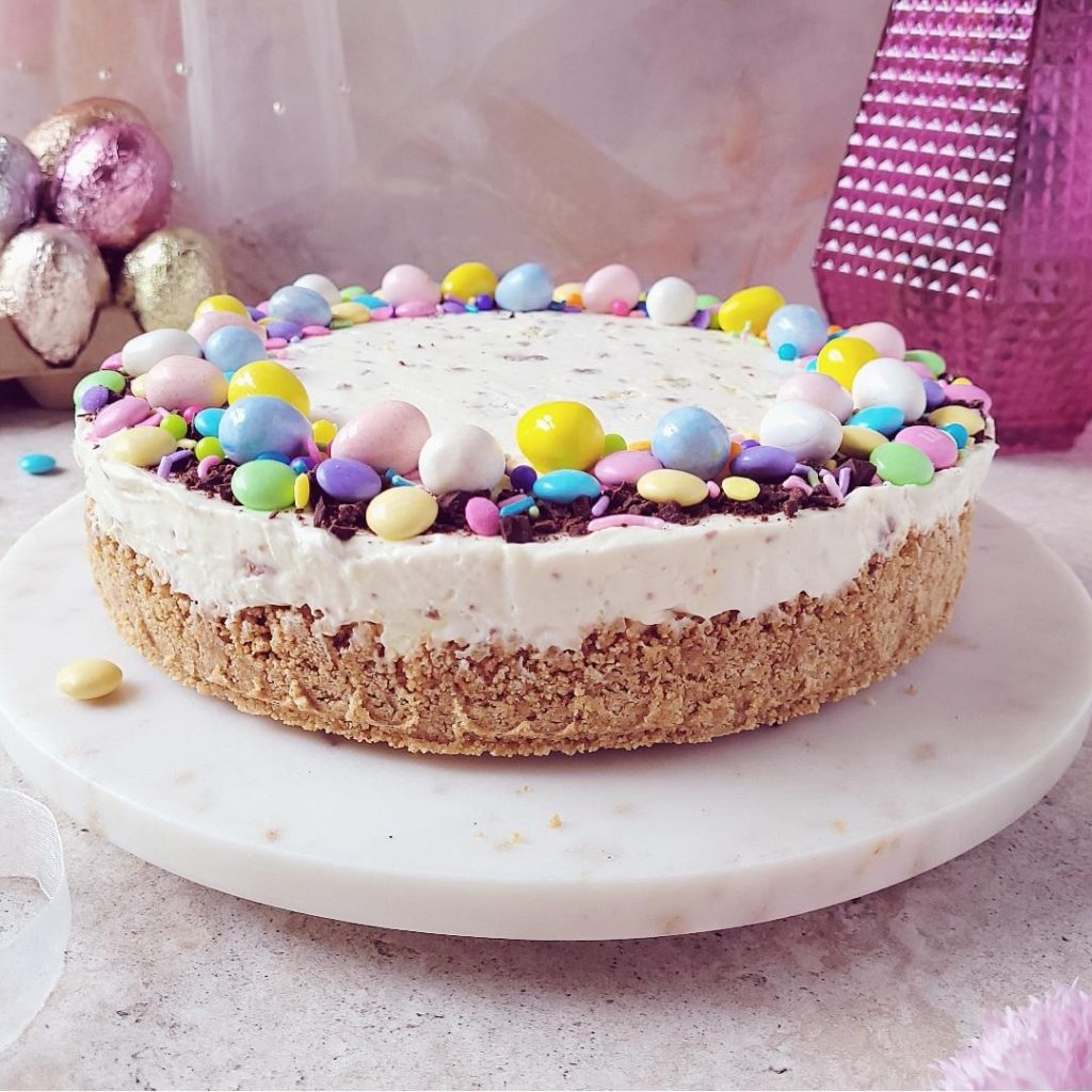 no bake easter cheesecake side view uncut decorated with mini eggs and easter candies pink background