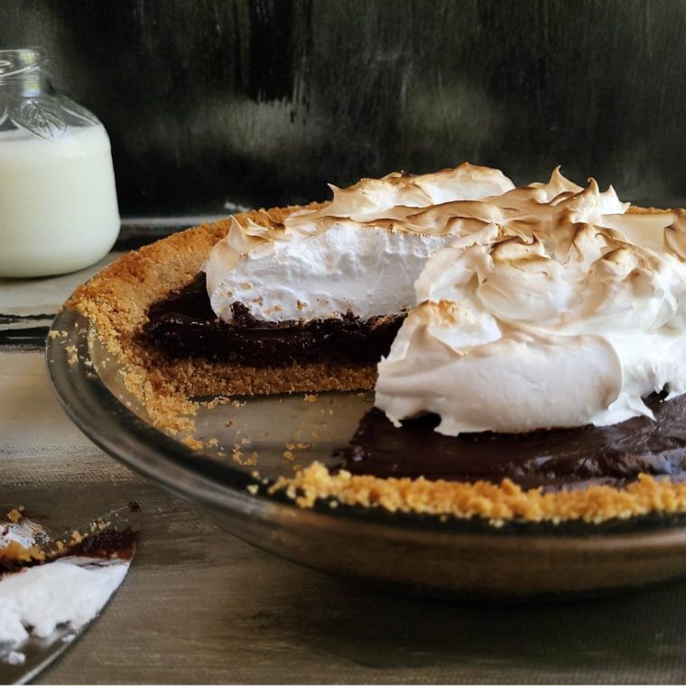 functional image chocolate smores pie in a glass pie dish with one slice missing to see layers of graham cracker crust, chocolate pie and toasted marshmallow topping black background