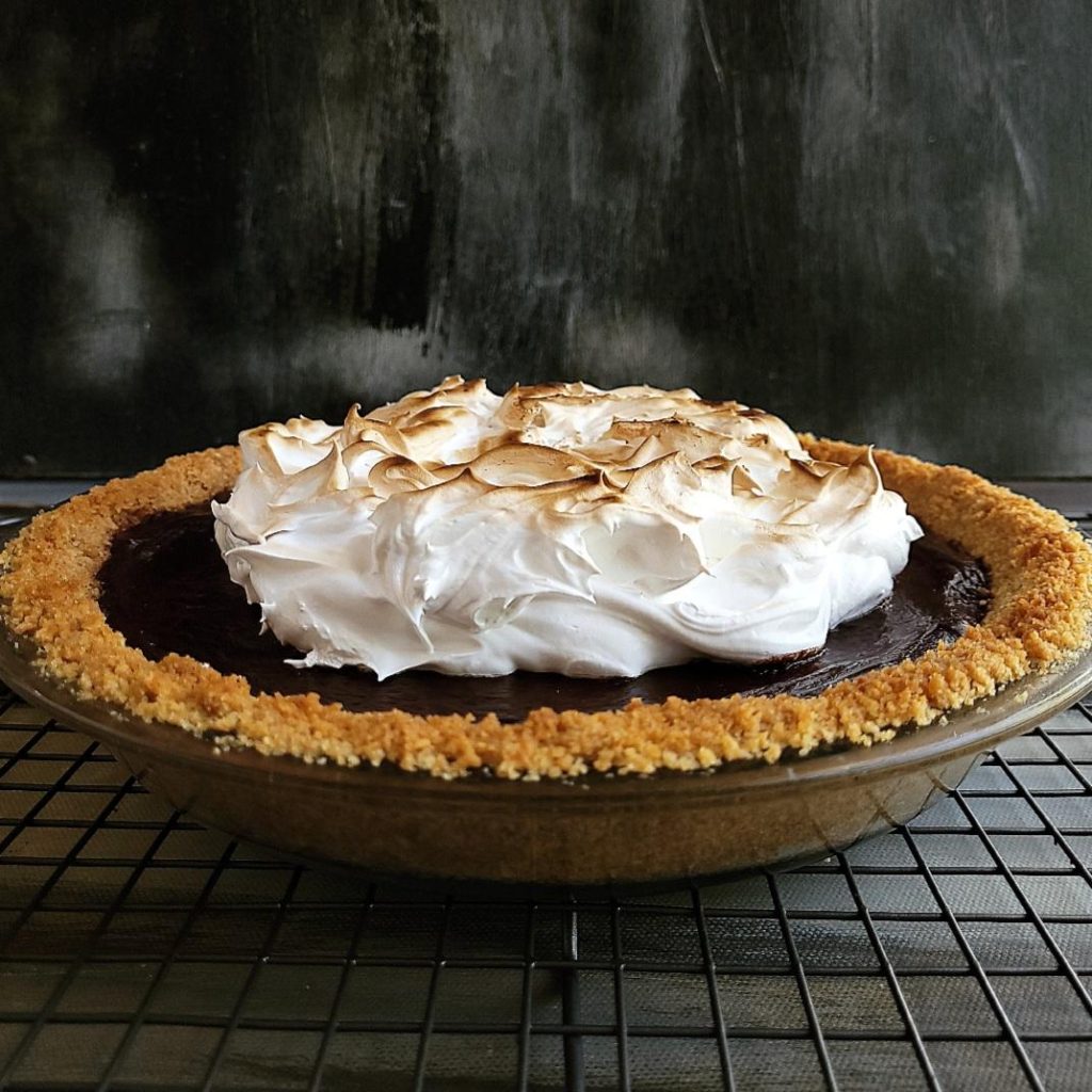 functional image chocolate smores pie uncut in pie dish on a black wire rack side view to see pile of toasted marshmallow topping on chocolate pie with graham cracker crust