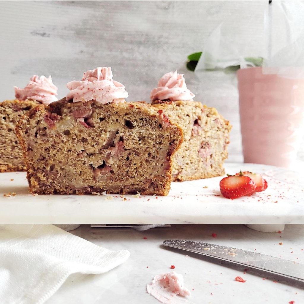 functional image strawberry banana bread three slices cut slices with strawberry frosting piped on top. styled on white marble with a pink vase of orchids in the background